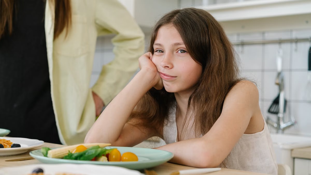 A young girl looking bored, sitting at a kitchen table with breakfast plates in front of her.