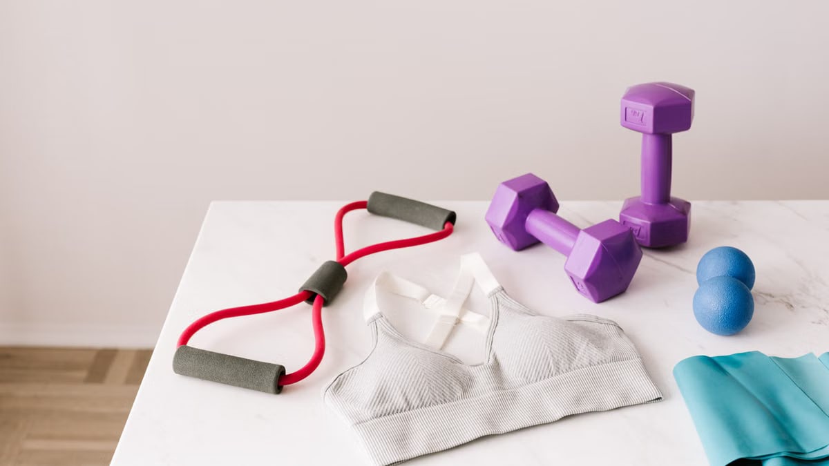 From above of white sports bra surrounded by purple dumbbells and blue tape expander and red tubular expander and blue massage double ball placed on marble surface of table